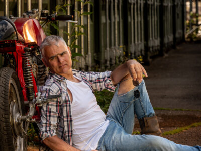 Stuart James sitting on the concrete ground while leaning against his classic red motorbike while his hand rests on leg, holding his sunglasses with a few green boxes behind it.